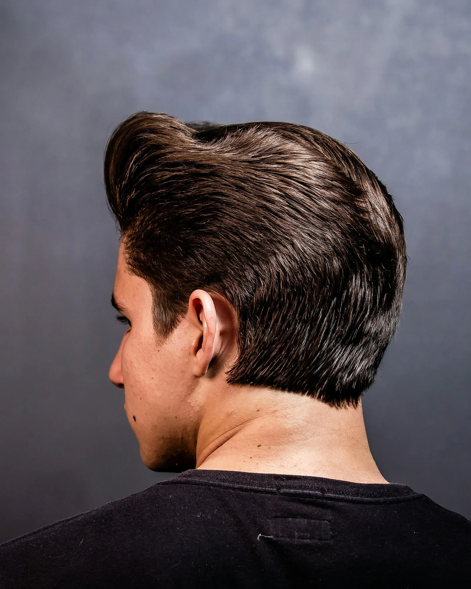 Vista posterior de un hombre con corte clásico hacia atrás, cabello sano y peinado con volumen sobre fondo neutro.